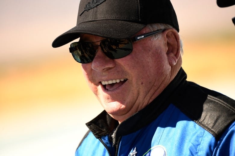 Willian Shatner wearing his flight outfit, black ball cap and sunglasses laughs as he looks off past the camera.