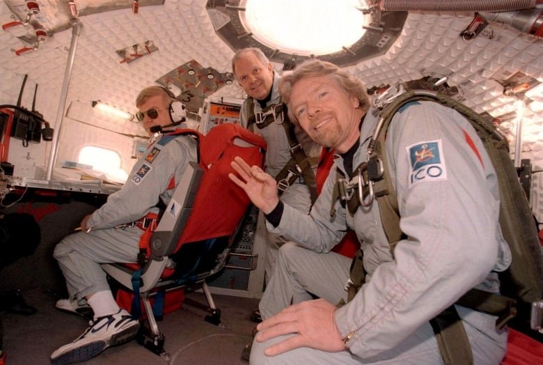 Richard Branson waves to the camera inside a capsule with two men behind him.