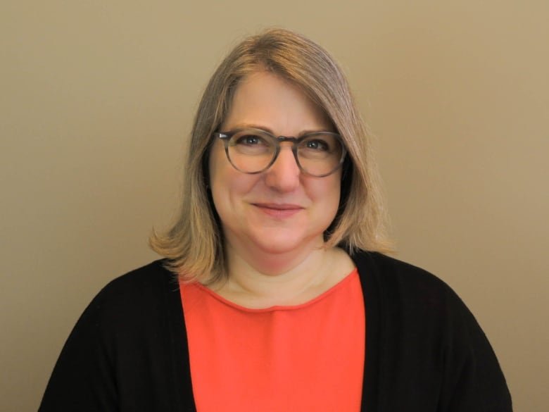 A woman with a short blonde bob wearing a orange shirt and black sweater cardigan is smiling at the camera.