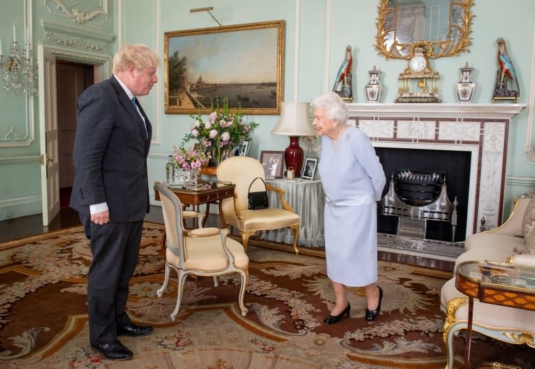 Two people stand while speaking with one another in a large formal sitting room.