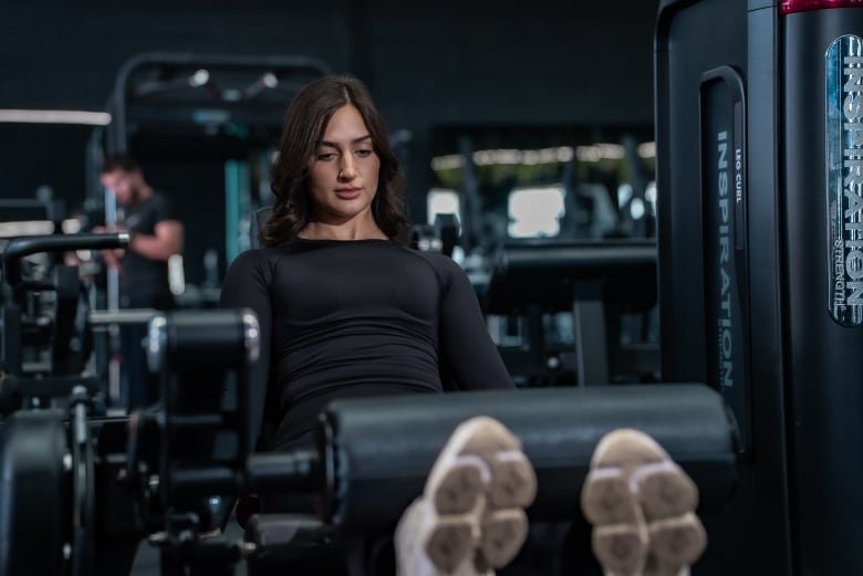 A young woman with black hair uses a leg workout machine at a gym. She is wearing sleek black workout clothing.