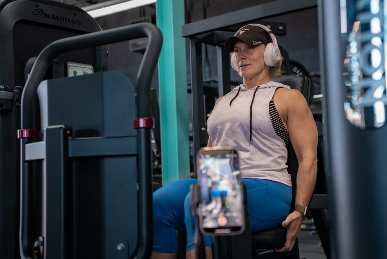 A woman wearing a baseball cap sits on an abductor gym machine. She is wearing a white workout tank top and blue leggings. There is a tripod with a phone camera recording her in front of her.