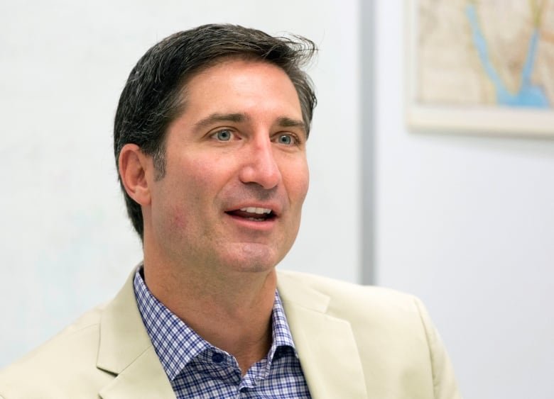 A cleanshaven man wearing a light-colored blazer and a collared checked shirt is shown in closeup speaking.