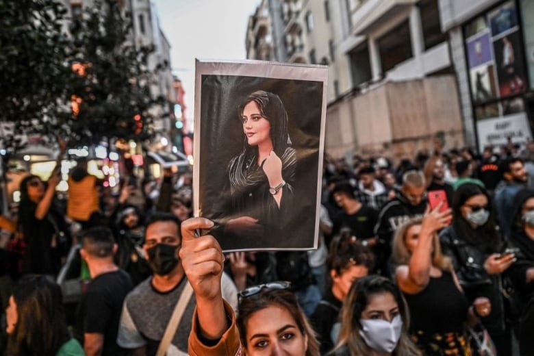 A woman in a crowd of protesters holds up a side profile picture of a woman wearing black clothing and a black hijab.