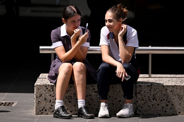 Two girls who appear to be teens are shown sitting on a step, with one holding up a phone.