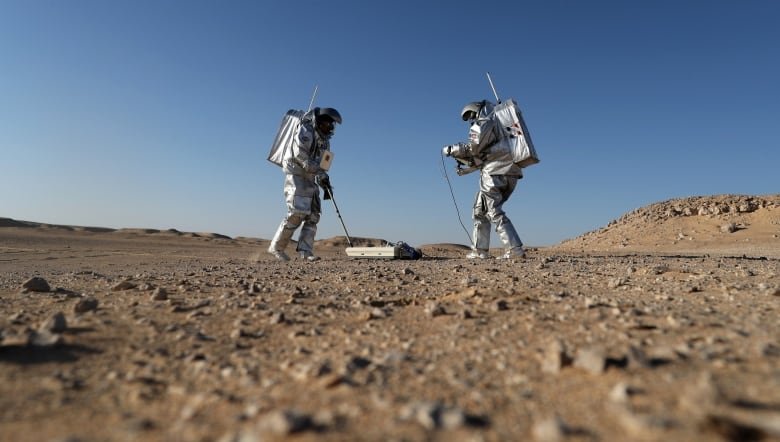 Two people in spacesuits stand in a desert with scientific equipment.