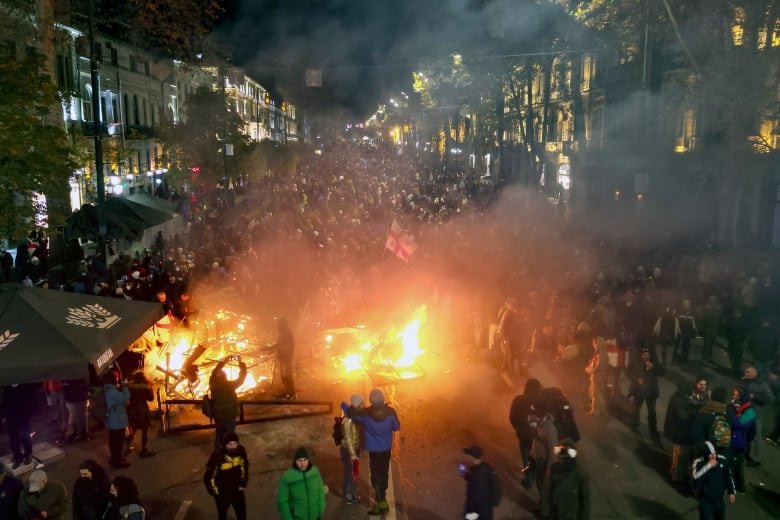 From a distance and a slight elevation, a nighttime street scene is shown in which hundreds of people are gathered in a protest. A fire has been set in the middle of the street, and a flag is seen raised in the air.