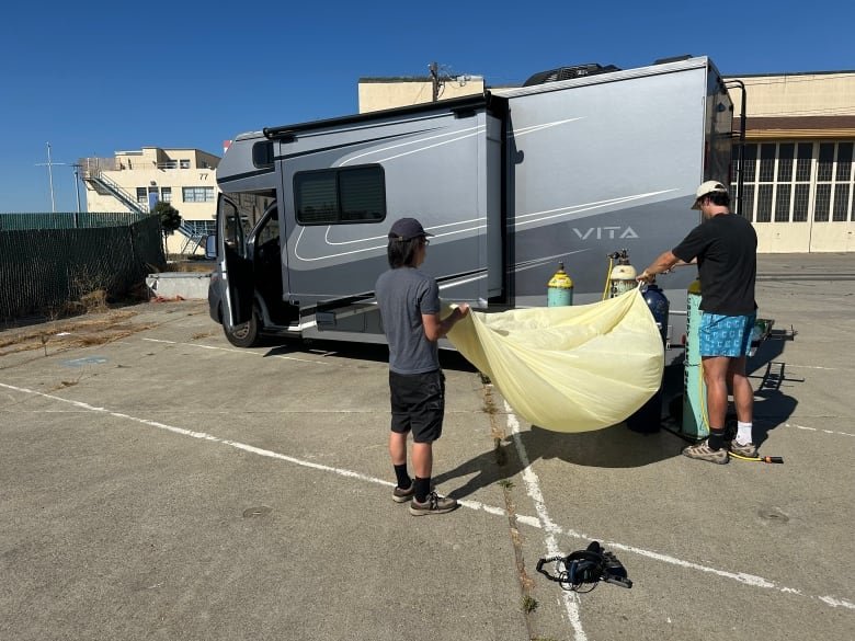 Two men stand outside a camper van while filling a balloon.