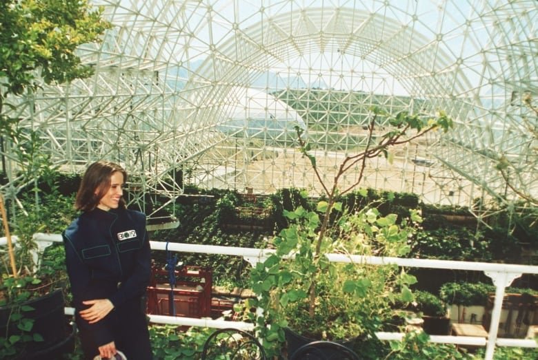 A woman stands, smiling, on a walkway in a building with a glass roof, greenery fills the massive room behind her.