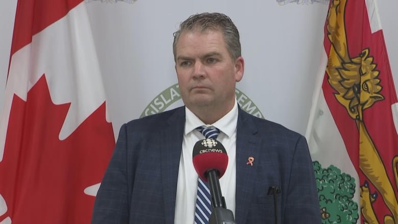 Bloyce Thompson stands against a backdrop of flags. He is wearing a white shirt, striped tie and black suit jacket. He stands in front of a CBC branded micorphone.