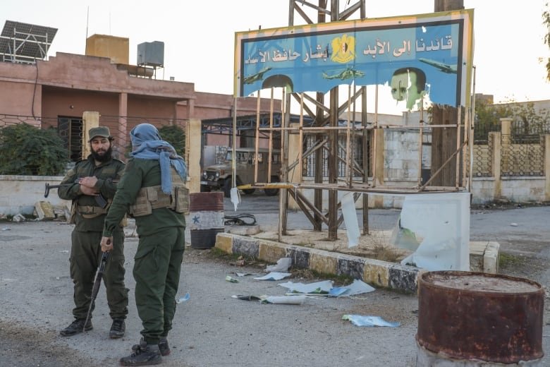 Two soldiers appear to be in conversation as they stand, with weapons, in a urban area underneath a ripped billboard.