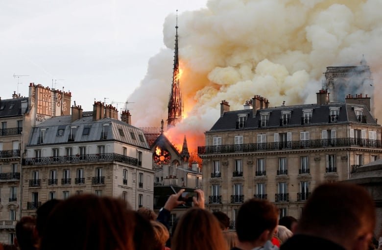 August buildings are shown, with orange fire surrounding a spire of a church. In the foreground are the backs of people's heads, as they look at the fire.