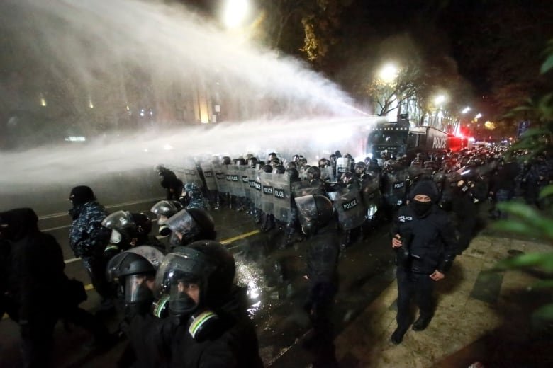 Several police officers are shown on the street in a nighttime scene, with massive sprays of water beside them.