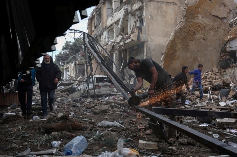 People walk on the rubble of damaged buildings as a man cuts steel.