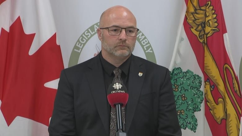 Robin Crouchers stands against a backdrop of flags in an all black suit.