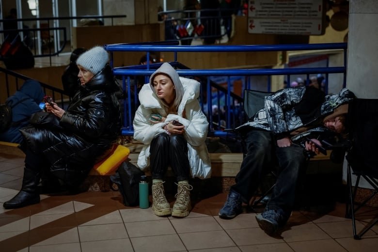 Three women in winter coats sit near a railing in a metro station.