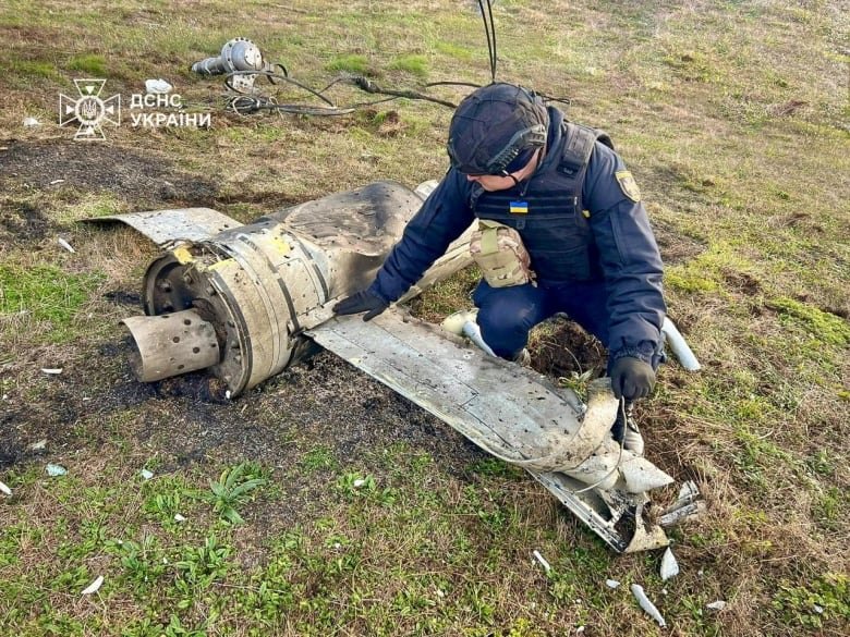 A man with a helmet is bent over the ground, touching a piece of metal that's laying on grass.