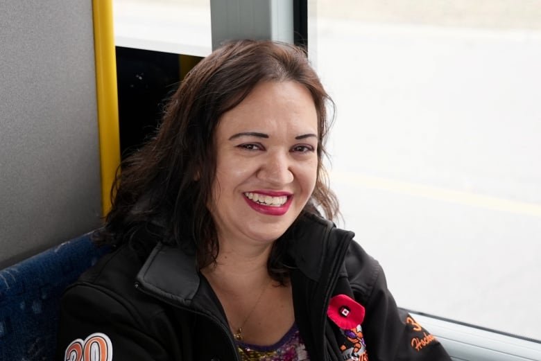 A woman with shoulder length dark hair, red lipstick and a dark jacket sits in a bus set by the window smiling at the camera.