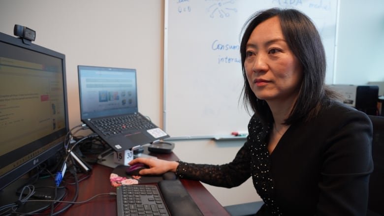 A woman works at a desk with two screen and two keyboards. There is a whiteboard in the background.