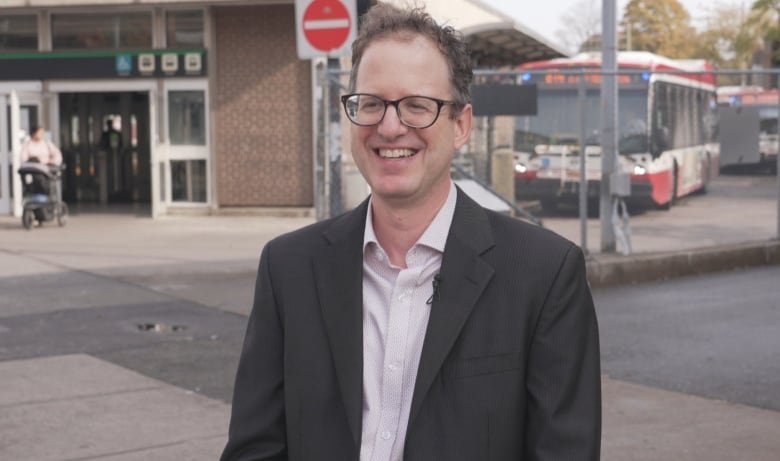 A man with short brown hair and glasseswearing a grey suit jacket stands in front of TTC station smiling.