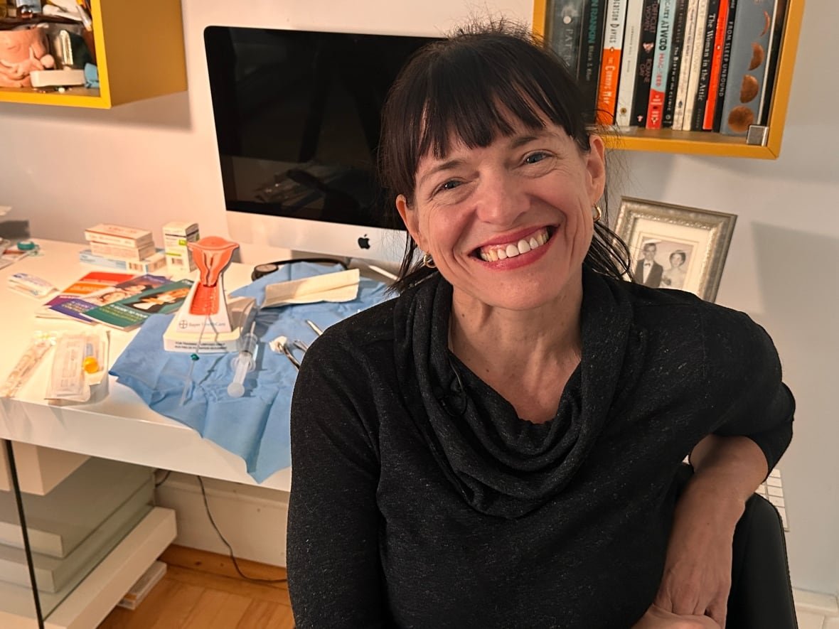 dr-kelly-monaghan A woman with dark hair and bangs sits in front of a model uterus and medical tools on a table.
