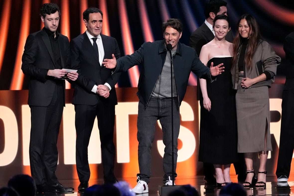 A person gestures while speaking on stage as others surround them.