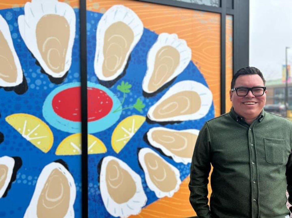 dylan-rose A person wearing thick, black glasses stands smiling in front of an art piece.