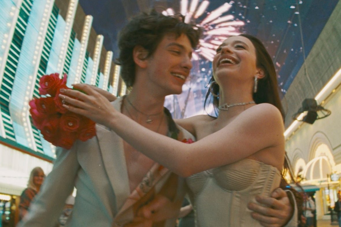 A smiling man and woman embrace. Behind them are neon lights and fireworks.