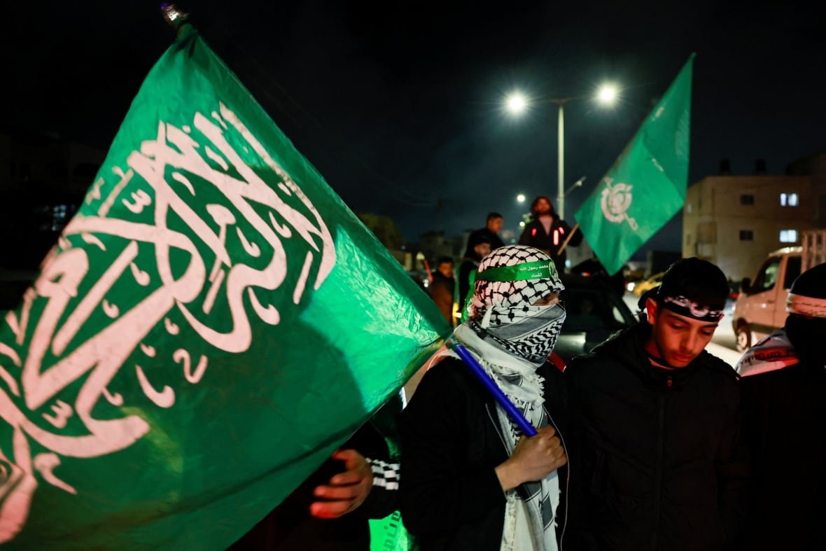israel-palestinians-gaza-ceasefire A masked man moves through a large crowd at night, carrying a large green flag.