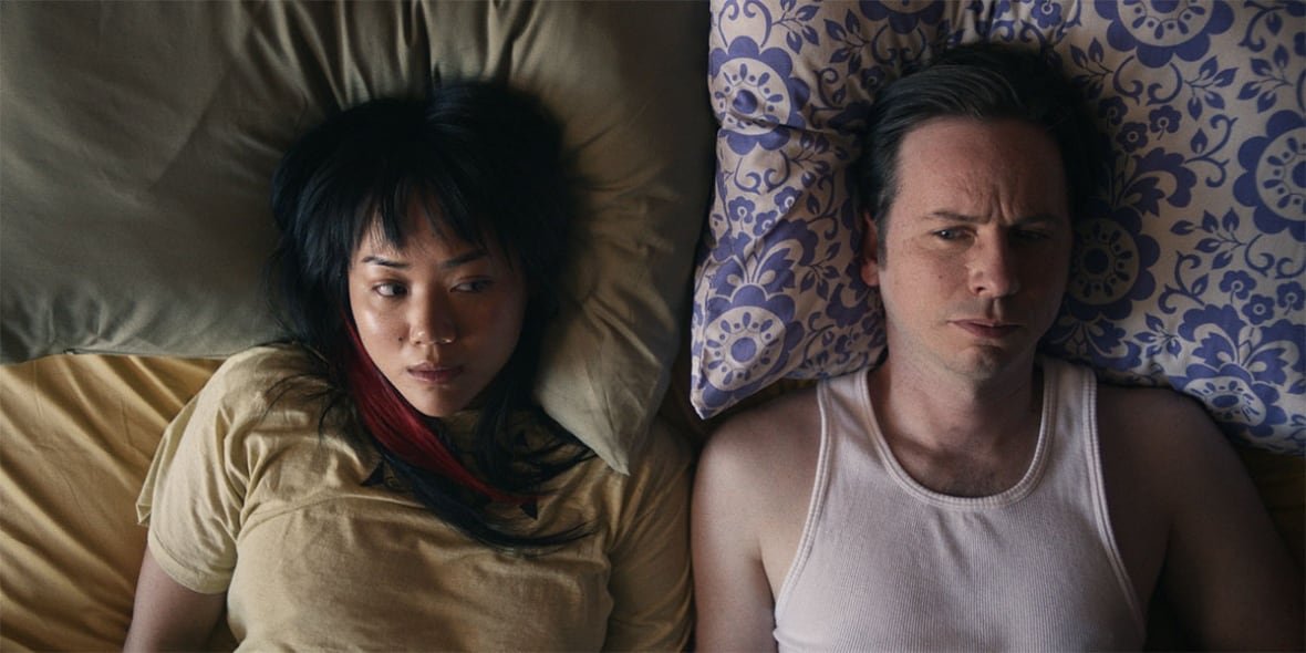 paying-for-it A man and woman lie next to each other in bed, eyeing each other sideways.