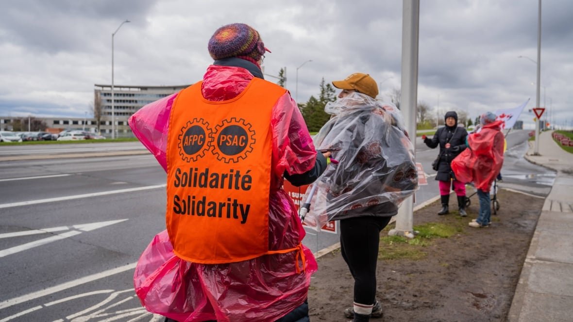 strike-psac-cra-canada-revenue-agency-picket-line-heron-road-ottawa Several people stand in a line on the side of a busy street in the rain.