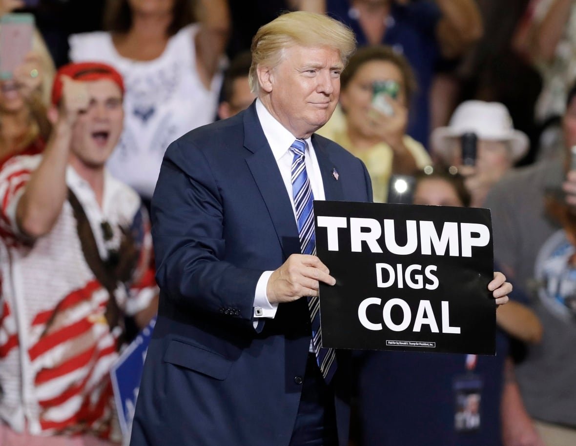 trump-digs-coal A man in his 80s with blond hair holds a black sign with white bold letters that says 'Trump Digs Coal.'