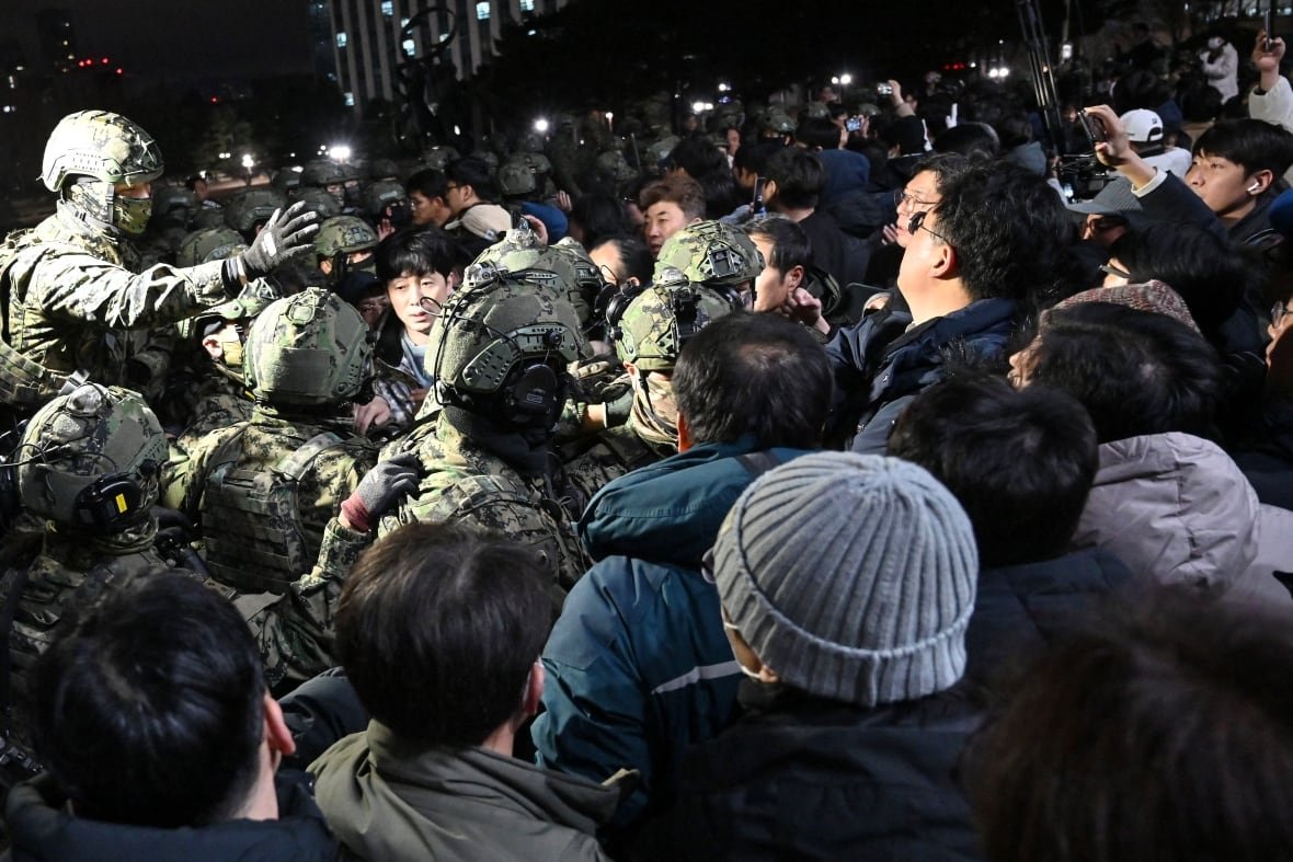 2187503273 A light shines on a mass of Asian people in a nighttime people, with soldiers and civilians jostling for position.