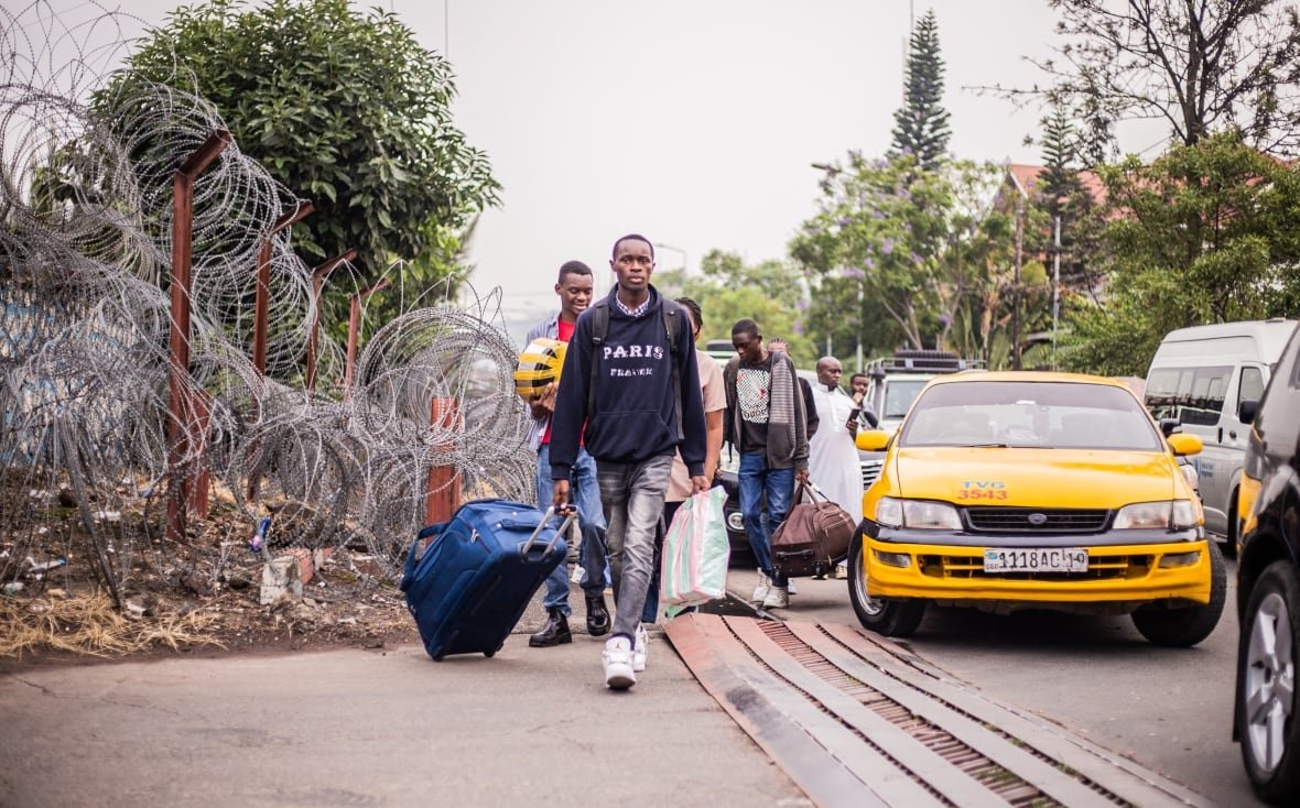 2195237967 People carry their belongings as they walk along a sidewalk bordered by barbed wire.
