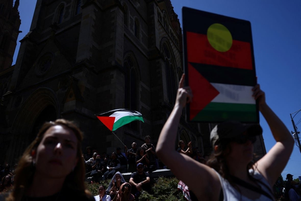australia-day-protests-1 A person carries a sign with the Australian Aboriginal flag above a Palestine flag in a crowd of protesters.
