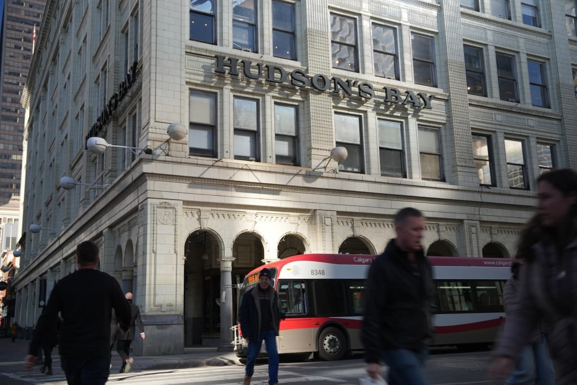 calgary-hudson-s-bay-1 A bus blurs past a building that says "Hudson's Bay" on it in black 3d letters. Several blurry silhouettes of people walk on the street in front of it.
