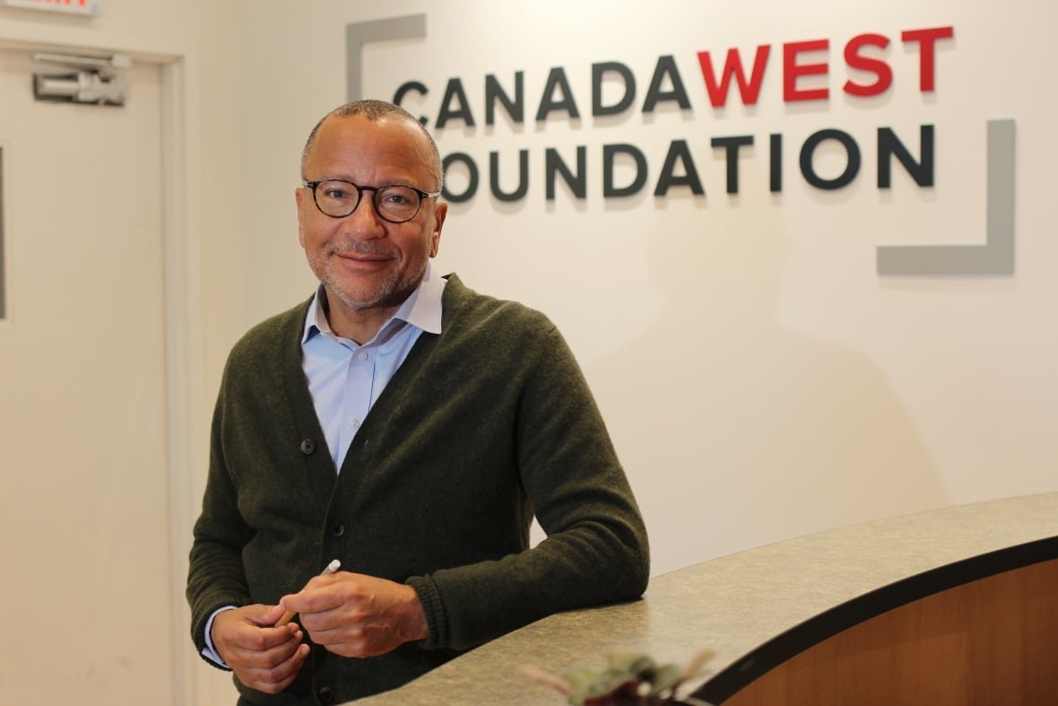 carlo-dade A man wearing glasses, a blue button-down shirt and green cardigan stands in front of a wall bearing the logo of the Canada West Foundation.