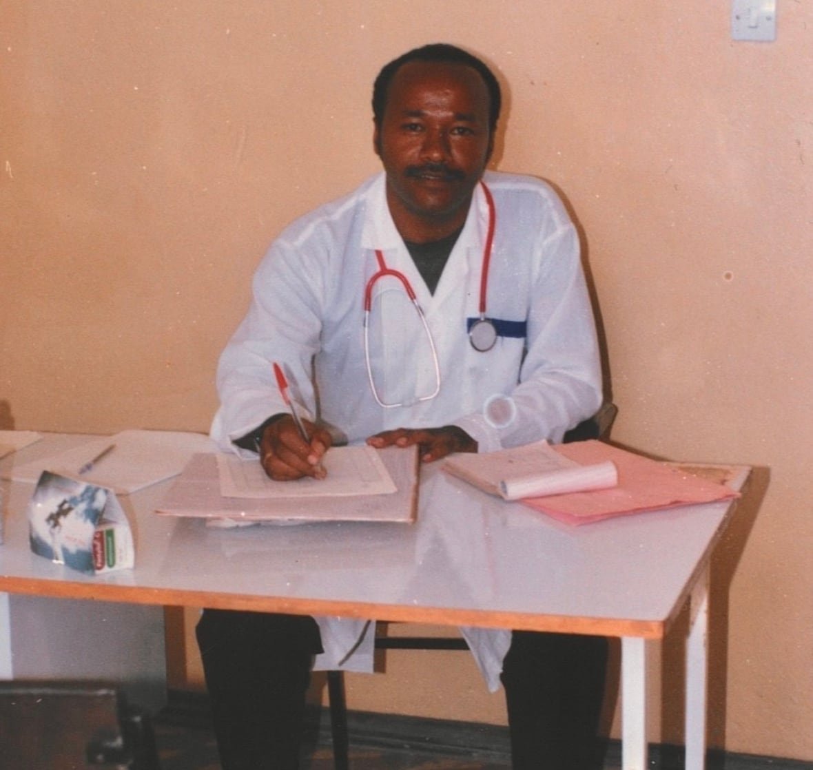 dawit-yakob-first-person A smiling man wearing a white lab coat and a stethoscope around his neck sits at a table doing paperwork.