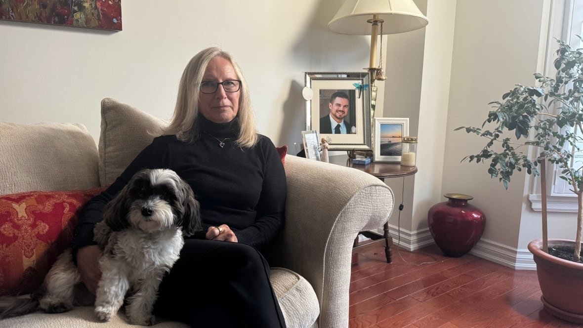 denise-turner A person sits on a couch with a dog beside them. A picture of a person is on a table behind them.