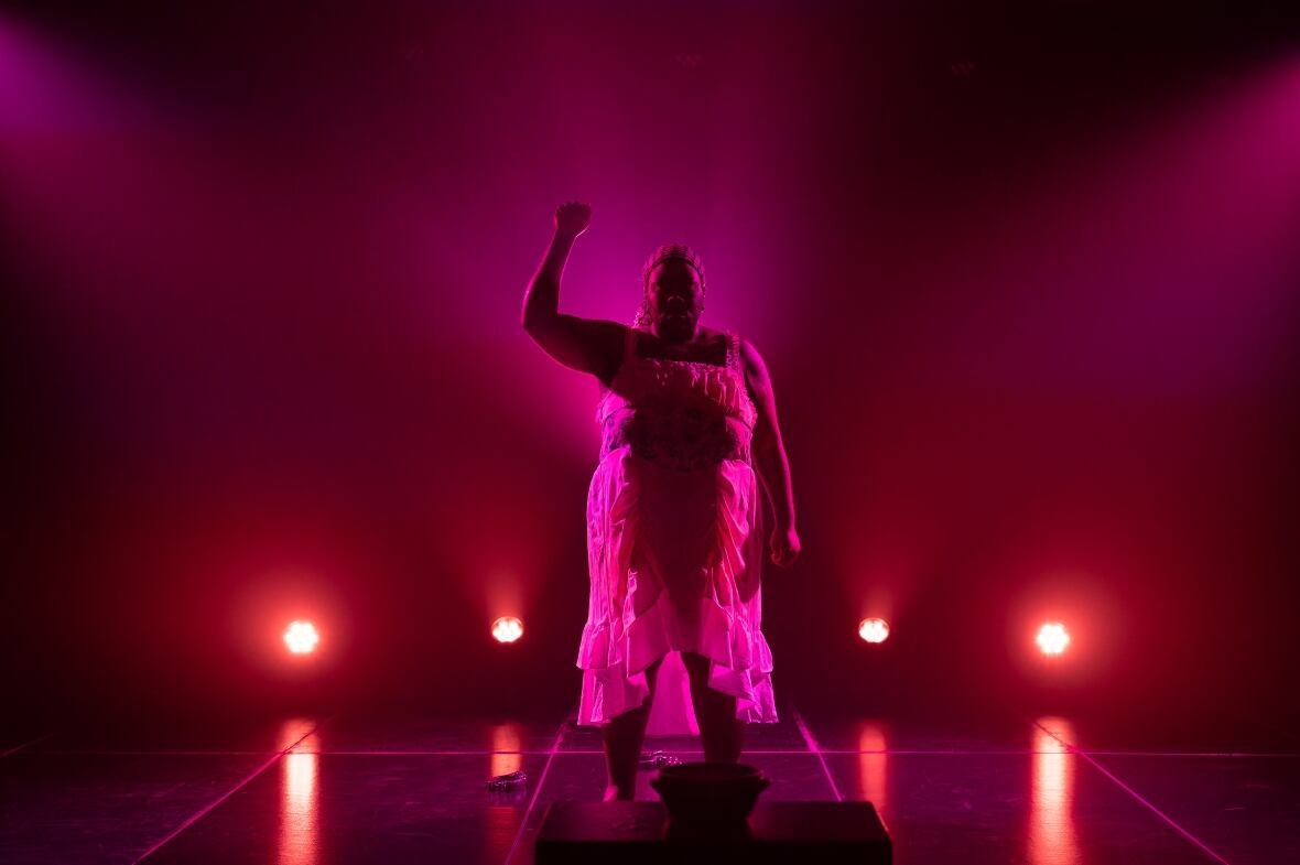 dragcessible-envy-the-clown Red and purple lighting light up a stage where a performer in a white dress holds up their right fist in the air.