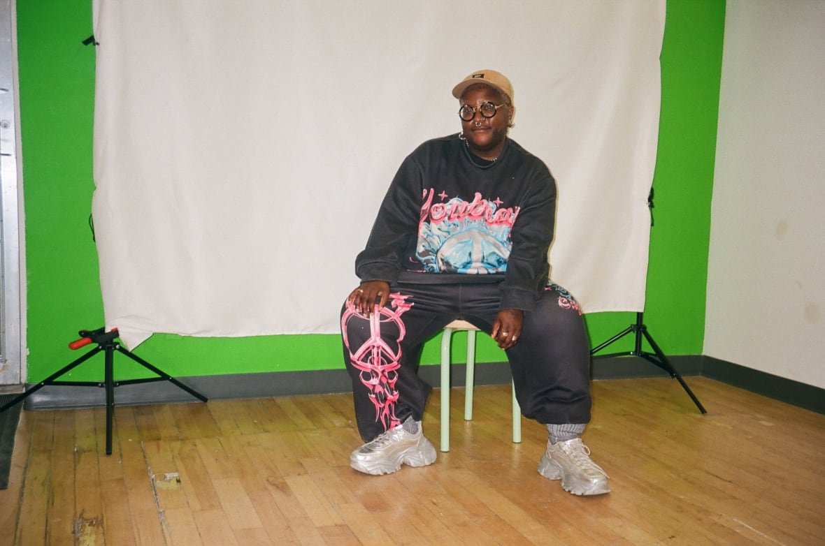 frankieb-lambert A Black person wearing a black and pink tracksuit with silver running shoes sits in front of a white screen in a studio.