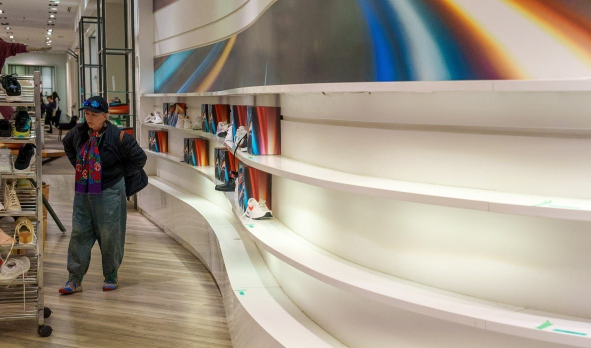 hudson-s-bay-liquidation-3 A person stands with their hands on their hips, looking at racks of shoes, at a department store interior. A curvy wall with shelves displaying shoes is seen at the right side of the image, mostly empty except for a few pairs of shoes.