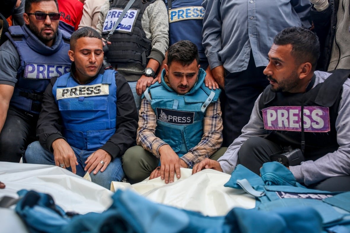 israel-gaza-28 Four men in press vests kneel in front of bodies covered in white and laid out on the ground.