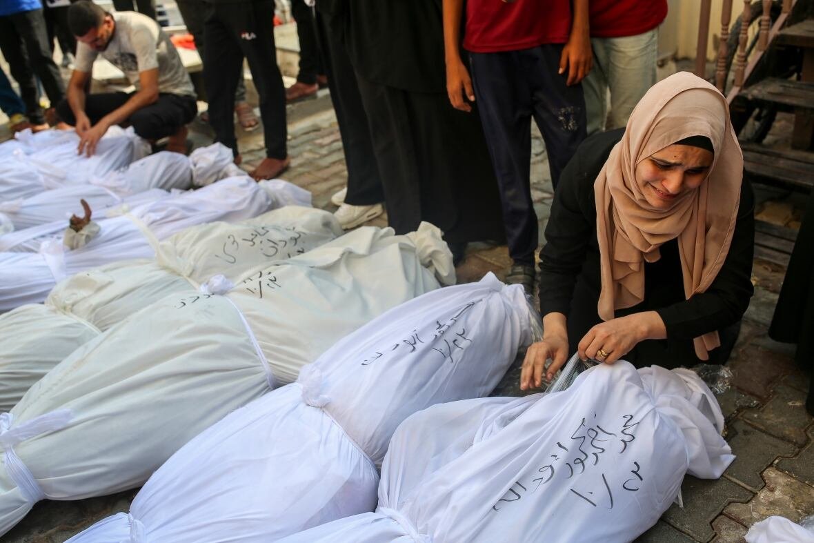 israel-gaza-29 A woman in a headscarf kneels on the ground sobbing next to a row of bodies, wrapped in white cloth, and laid out next to each other.