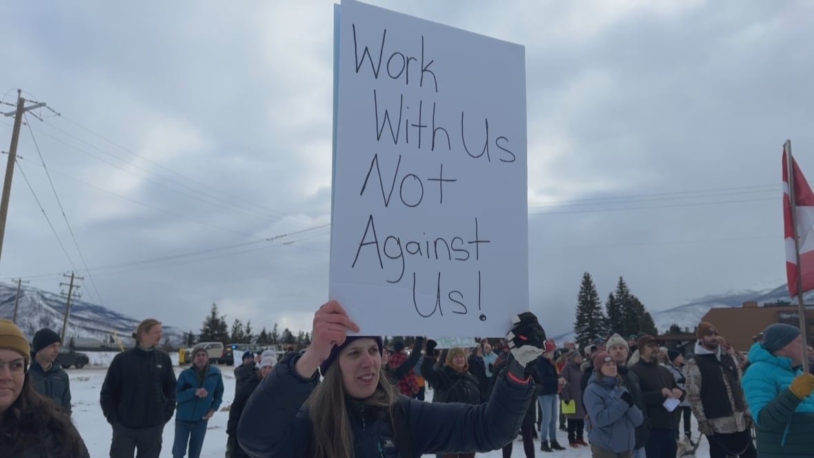 jasper-housing-rally-1 A woman, bundled in winter clothes outside, is holding up a sign that says, 'Work with us, not against us!' She is standing amongst a crowd of people, under a cloudy sky.
