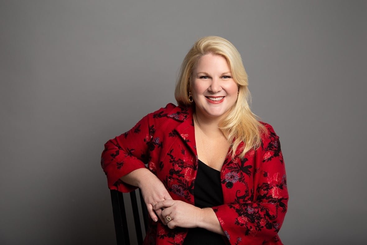laura-tamblyn-watts Smiling blonde woman, wearing a red blazer with a floral pattern, poses in a chair in front of a grey studio backdrop.