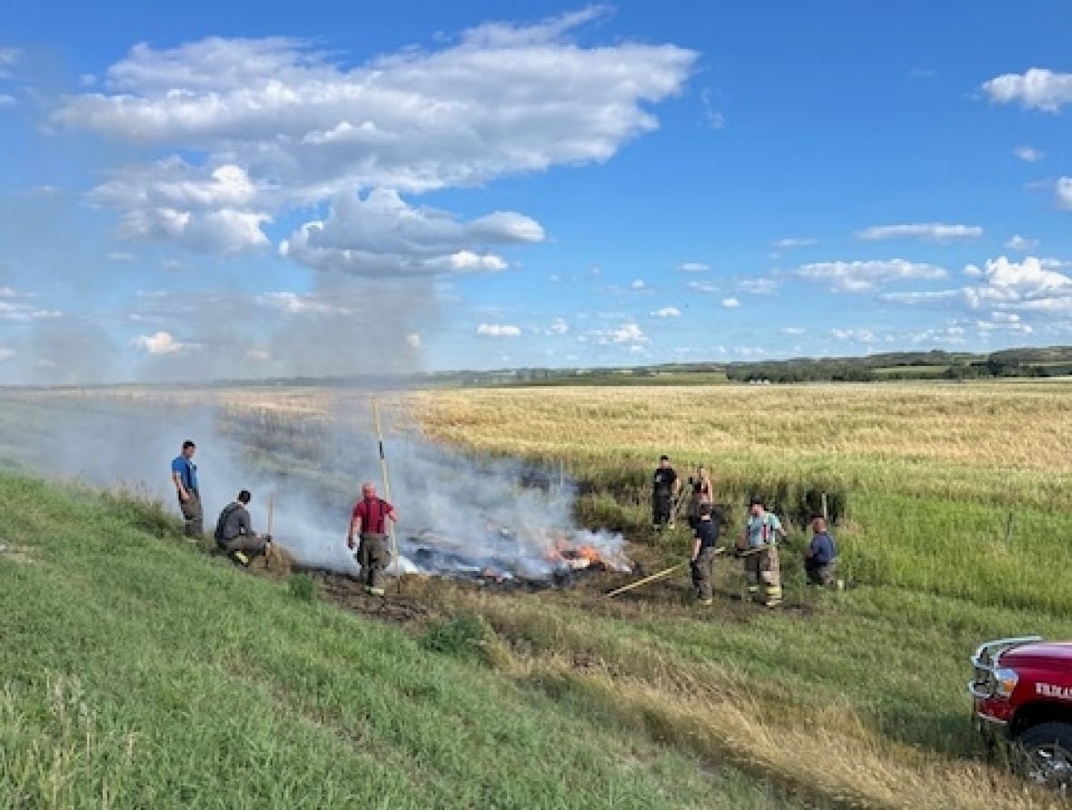 macklin-hay-bale-arsonist People with tools stand around a burning bale in a field during the day.