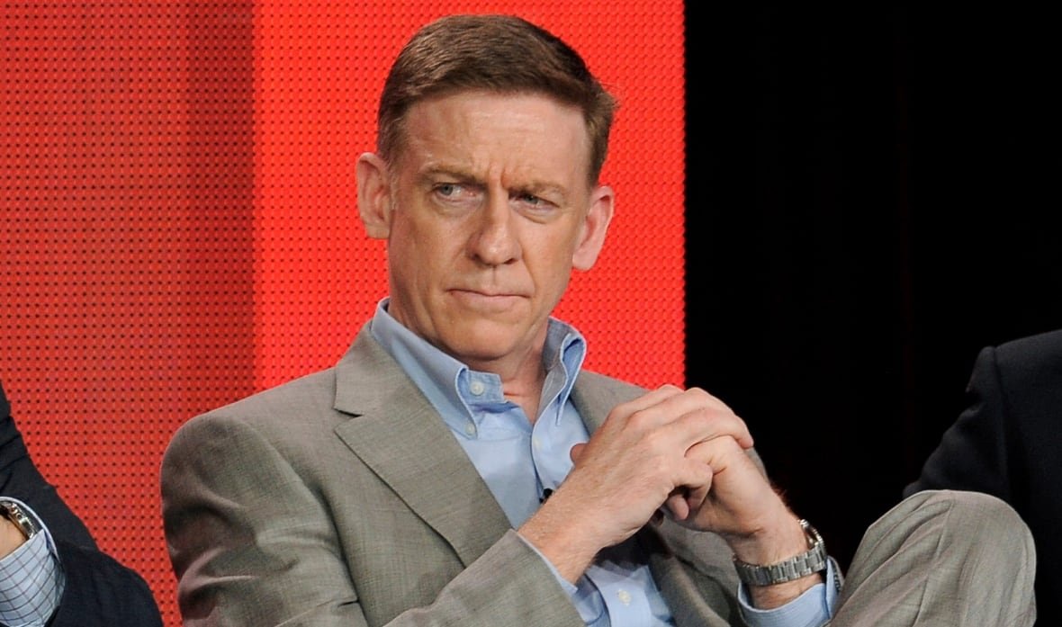 media-cbs-60-minutes A man in a beige suit and a blue shirt sits in a chair with a serious expression and his hands folded in front of his chest. He is watching something off camera and there is an orange backdrop behind him.