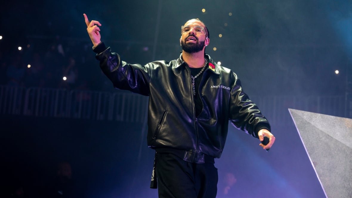 music-drake-tour-20241125 A man is shown from the knees up, walking with his hand in the air, pointing to the sky. He appears to be in the middle of speaking or singing, and there is a dark blue backdrop behind him.