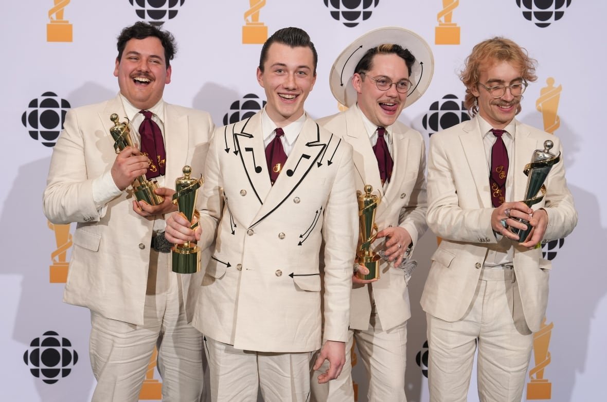 music-juno-awards-20250329-9 Dressed in matching retro suits with music note details on the collars, four musicians celebrating with their trophy after winning a big musical award.